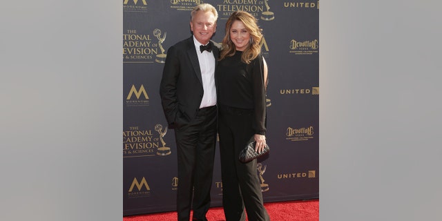 Pat Sajak in a black classic tuxedo smiles next to his wife with a black gown with slits on the sleeves