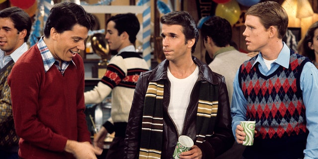 Anson Williams as Potsie, Henry Winkler as Fonzie, and Ron Howard as Richie hold cups at a party while getting something to drink in an episode of Happy Days, with Winkler wearing his character's signature leather jacket and a striped scarf