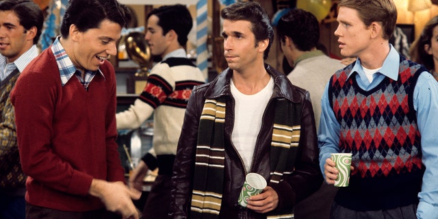 Anson Williams as Potsie, Henry Winkler as Fonzie, and Ron Howard as Richie hold cups at a party while getting something to drink in an episode of Happy Days, with Winkler wearing his character's signature leather jacket and a striped scarf