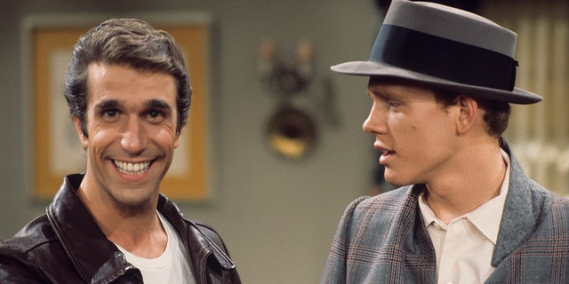 Henry Winkler as Fonzie stares at the camera and flashes a smile as Fonzie in a white shirt and leather jacket as Ron Howard as Richie stares at him wearing a brimmed hat in a season 3 episode of "Happy Days"