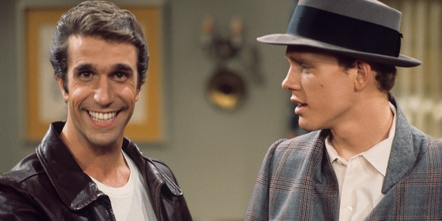 Henry Winkler as Fonzie stares at the camera and flashes a smile as Fonzie in a white shirt and leather jacket as Ron Howard as Richie stares at him wearing a brimmed hat in a season 3 episode of "Happy Days"