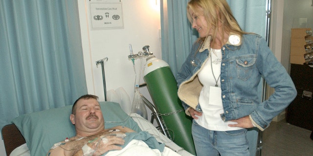 Bo Derek wearing a denim jacket and jeans with a matching white shirt speaking with a man lying on a hospital bed