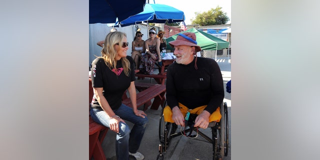 Bo Derek in a black outfit and sunglasses chatting with a man in a matching black outfit and a colorful hat