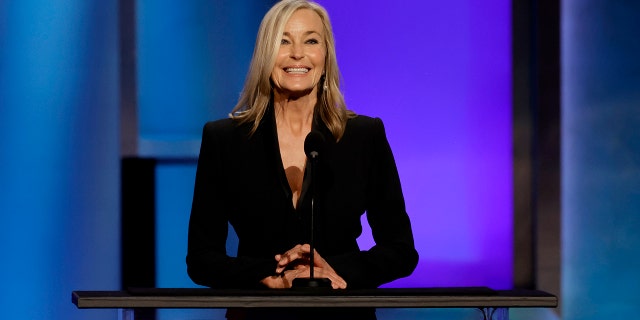 Bo Derek wearing black and smiling while standing in front of a podium