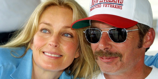 Bo Derek in a blue shirt leaning against a man with sunglasses and a red and white hat