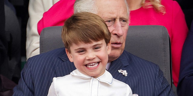 Prince Louis smiling and wearing a white shirt sitting on his grandfather king charles lap