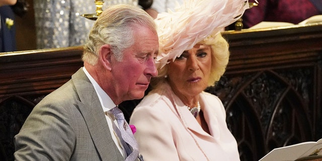Camillla wearing a pink dress and a matching hat sitting next to Charles in a grey suit