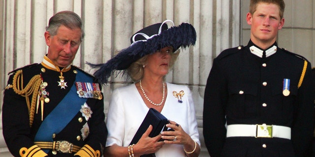 Prince Charles in royal regalia with Camilla wearing a white dress and an oversized navy hat looking at Prince Harry in uniform