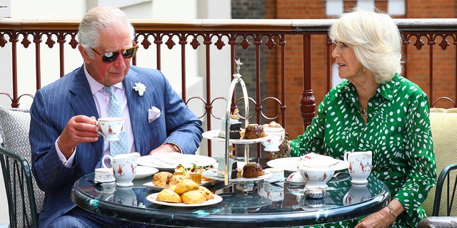 King Charles in a light blue suit sitting with Queen Camila in a green and white dress as they have tea