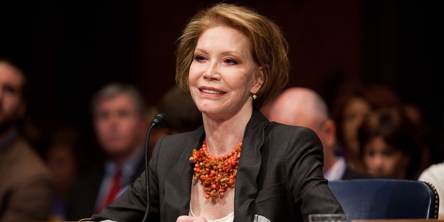 Mary Tyler Moore wearing a dark blazer and a white blouse with a coral necklace testifying