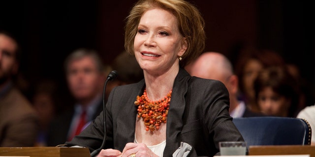 Mary Tyler Moore wearing a dark blazer and a white blouse with a coral necklace testifying