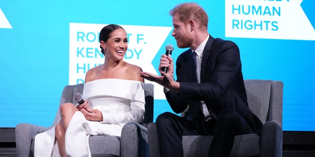 Meghan Markle wearing a white off the shoulder dress sitting next to Prince Harry in a suit and tie