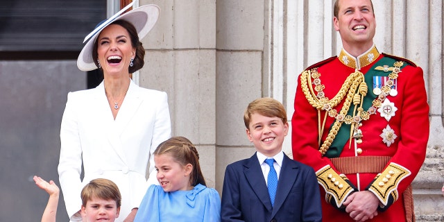 Kate Middleton wearing a white dress and a matching hat standing next to Prince William in a red royal uniform and their three children