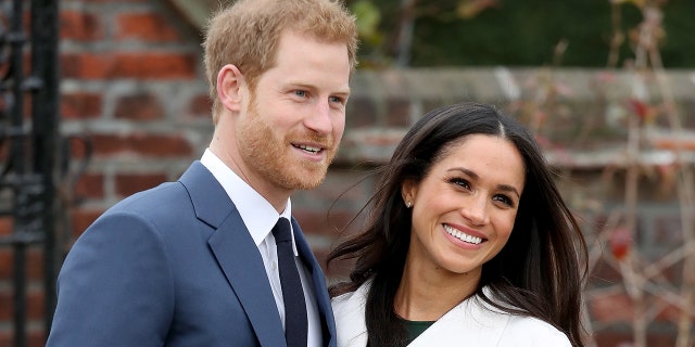 Meghan Markle wearing a white coat and a black dress next to Prince Harry in a blue suit and skinny black tie