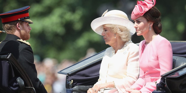 Prince Harry in his military uniform sitting across from Camilla in a white dress and matching hat and Kate Middleton in a pink dress and a matching hat