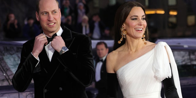 Prince William in a tux adjusting his bowtie walking next to Kate Middleton in a strapless white gown and black opera gloves