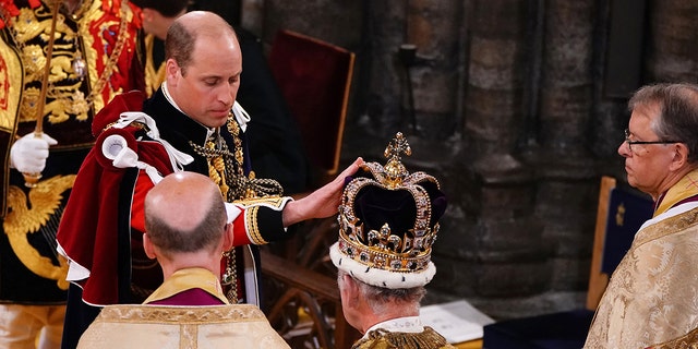 Prince William in royal regalia touching his father King Charles crown during the coronation ceremony