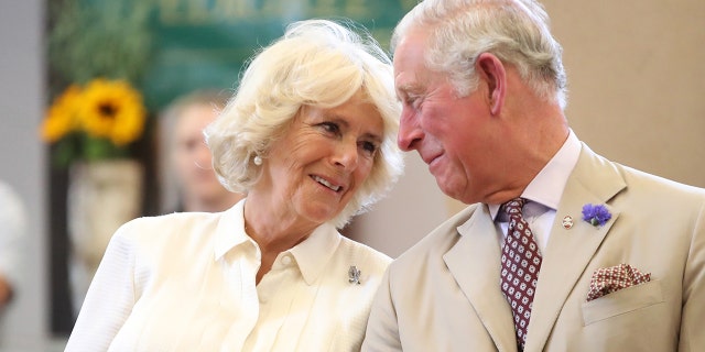 Queen Camilla in a beige blouse looks lovingly at her husband King Charles in a matching suit and tie