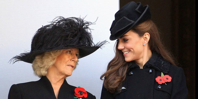Queen Camilla in a black dress and matching hat looking at a smiling Kate Middleton also wearing a black coat and matching dress