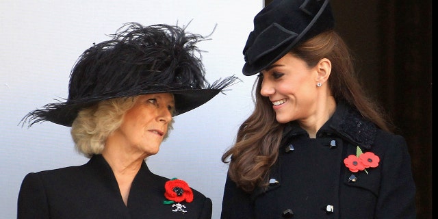 Queen Camilla in a black dress and matching hat looking at a smiling Kate Middleton also wearing a black coat and matching dress