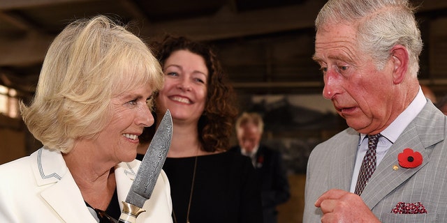 Camilla in a white blazer jokingly holding a knife as king charles in a grey suit looks on