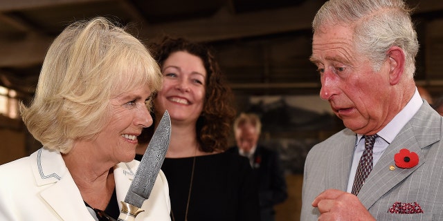 Camilla in a white blazer jokingly holding a knife as king charles in a grey suit looks on