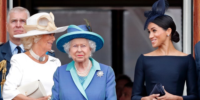 Camilla wearing a white dress chatting with Meghan Markle in a dark blue dress as Queen Elizabeth, wearing a multilayered blue dress, looks straighforward