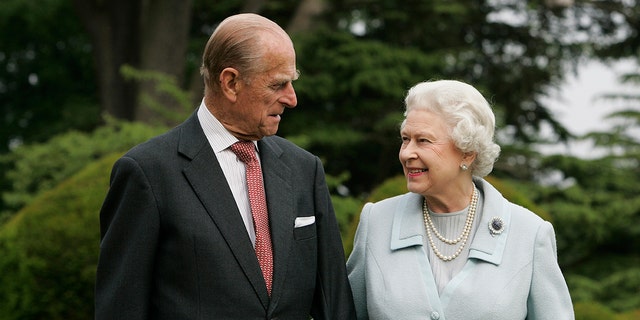 Prince Philip in a suit and tie looking at queen elizabeth in a blue dress and pearls