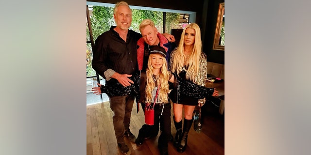 Eric Johnson in a black shirt and dark pants stands next to Joe Simpson, who is partially hunched wearing a pink furry jacket, next to Jessica Simpson in a mini skirt and sparkly gold top, and her daughter Maxwell Drew with a sparkly top in the front with her arms spread out