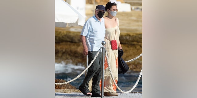 Robert De Niro wears a black cap and facemask and walks on a boardwalk in France with Tiffany Chen, in a long dress, also wearing a facemask