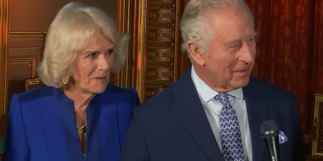 Queen Camilla in a royal blue suit, next to King Charles in a navy suit and blue patterned tie during an episode of "American Idol"