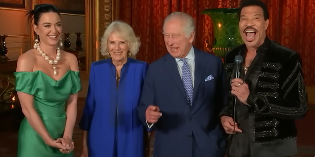 Katy Perry chuckles in a green gown with sleeves on the side and a massive pearl necklace next to Queen Camilla in a royal blue suit, next to King Charles in a navy suit and blue patterned tie next to Lionel Richie in black, holding a microphone