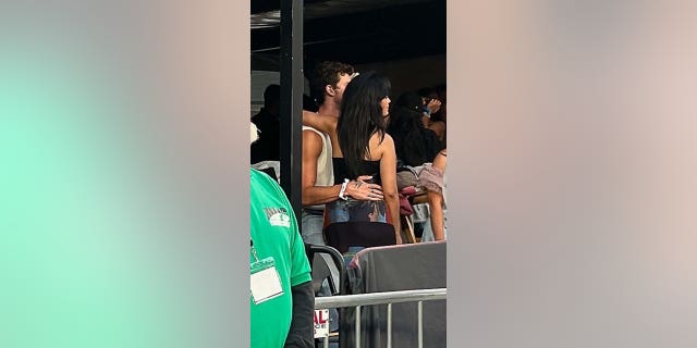 Camila Cabello in black wraps her arm around Shawn Mendes neck with his hand behind her back at the Taylor Swift Era's Tour