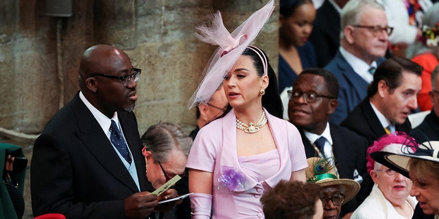 Katy Perry in a lilac dress and large headband hat struggles to find her seat in Westminster Abbey