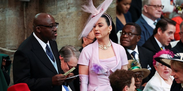 Katy Perry in a lilac dress and large headband hat struggles to find her seat in Westminster Abbey