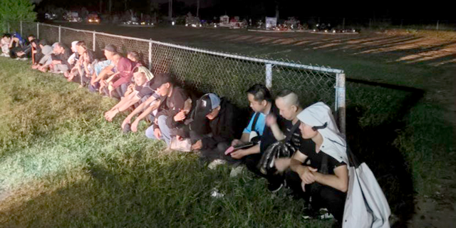 A line of migrants sat along a fence at night