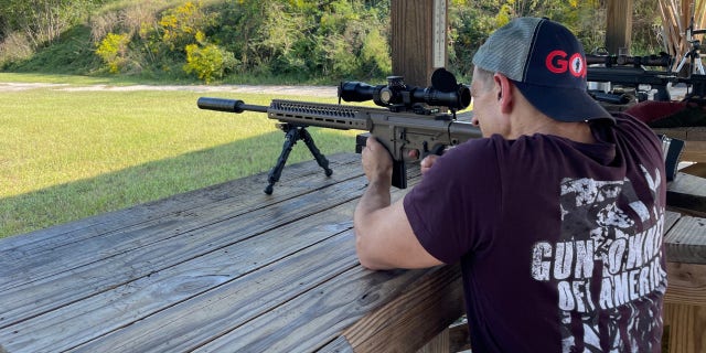 A man wearing Gun Owners of America merchandise shoots a rifle