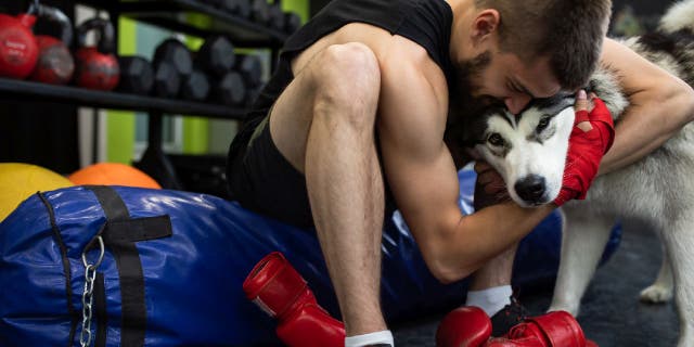 A young man is sitting on a punching bag in a gym hugging his dog.