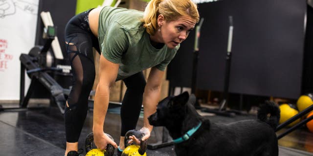 A woman deadlifts with kettlebells in a gym while her dog watches.
