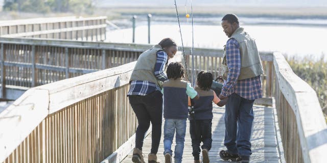 Rear view of a young family with two boys going fishing. They are walking on a wooden walkway or bridge, carrying rods and a tackle box.