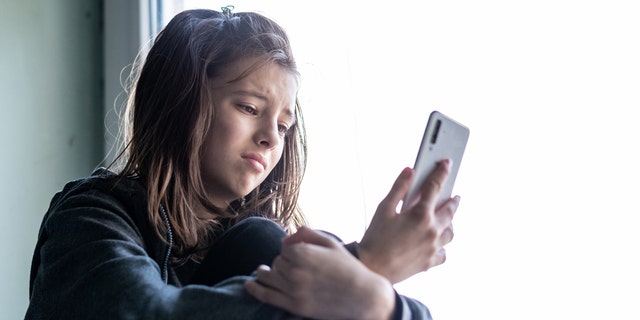 Teenage girl sitting at the window and looking at her phone with a sad expression