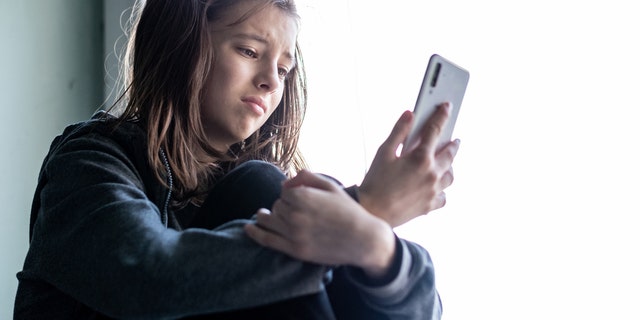 A teenage girl sitting at the window and looking at her phone with a sad expression