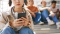 Closeup of teenage girl holding smartphone outdoors while sitting on metal stairs with group of friends in background, copy space