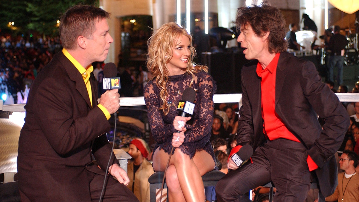 Kurt Loder interview Britney Spears in a black dress who looks at Mick Jagger in a red shirt and black suit