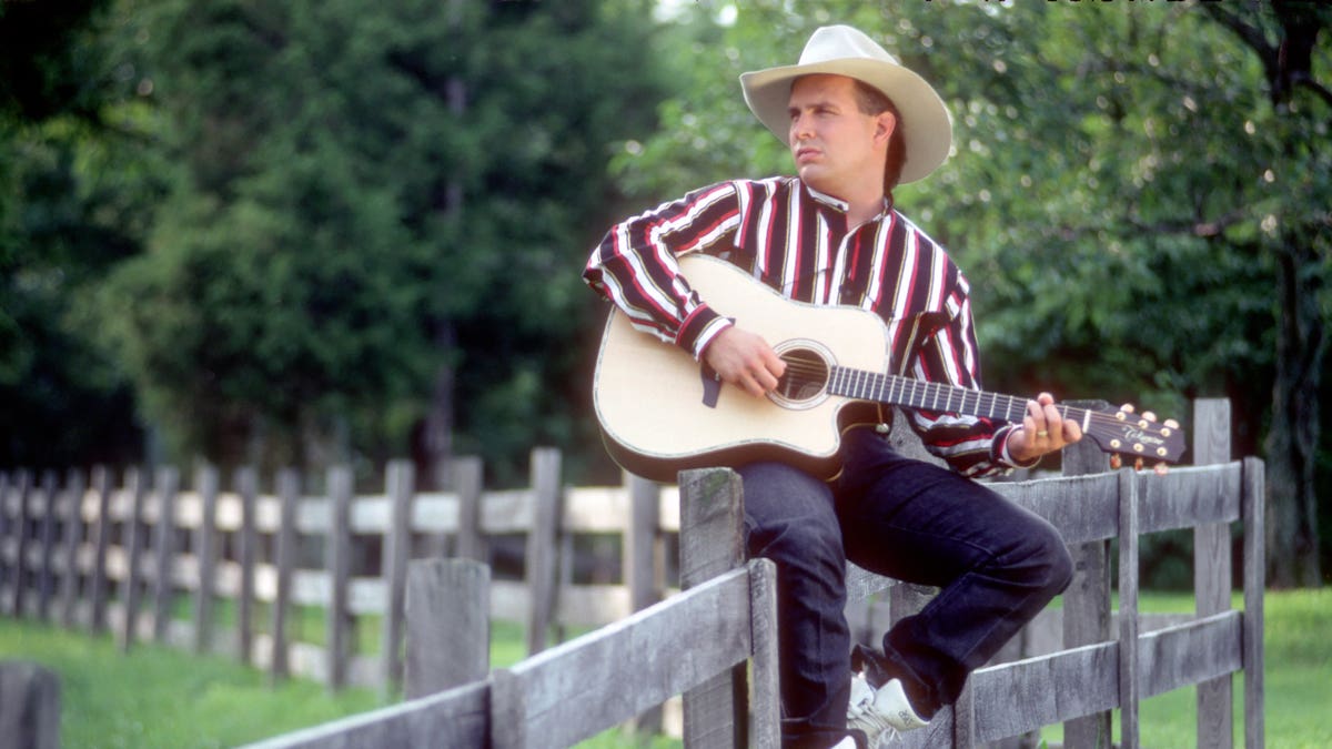 Garth Brooks sitting on a fence with a guitar wearing a cowboy hat.