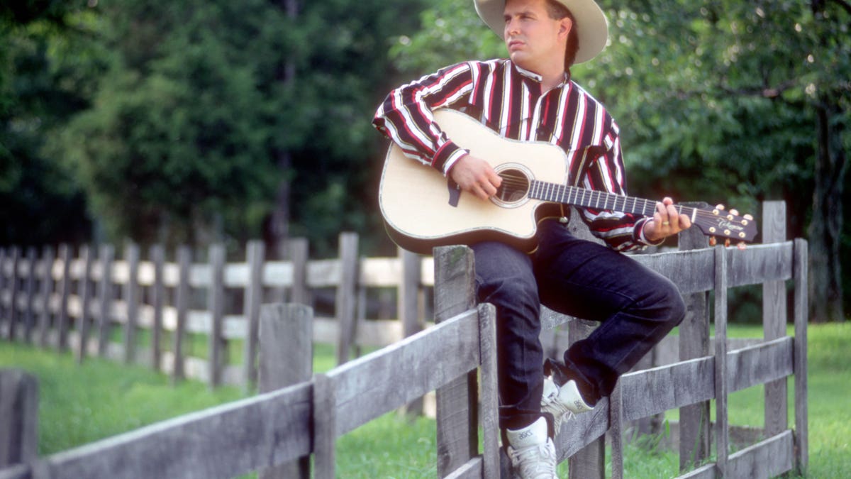 Garth Brooks sitting on a fence with a guitar wearing a cowboy hat.