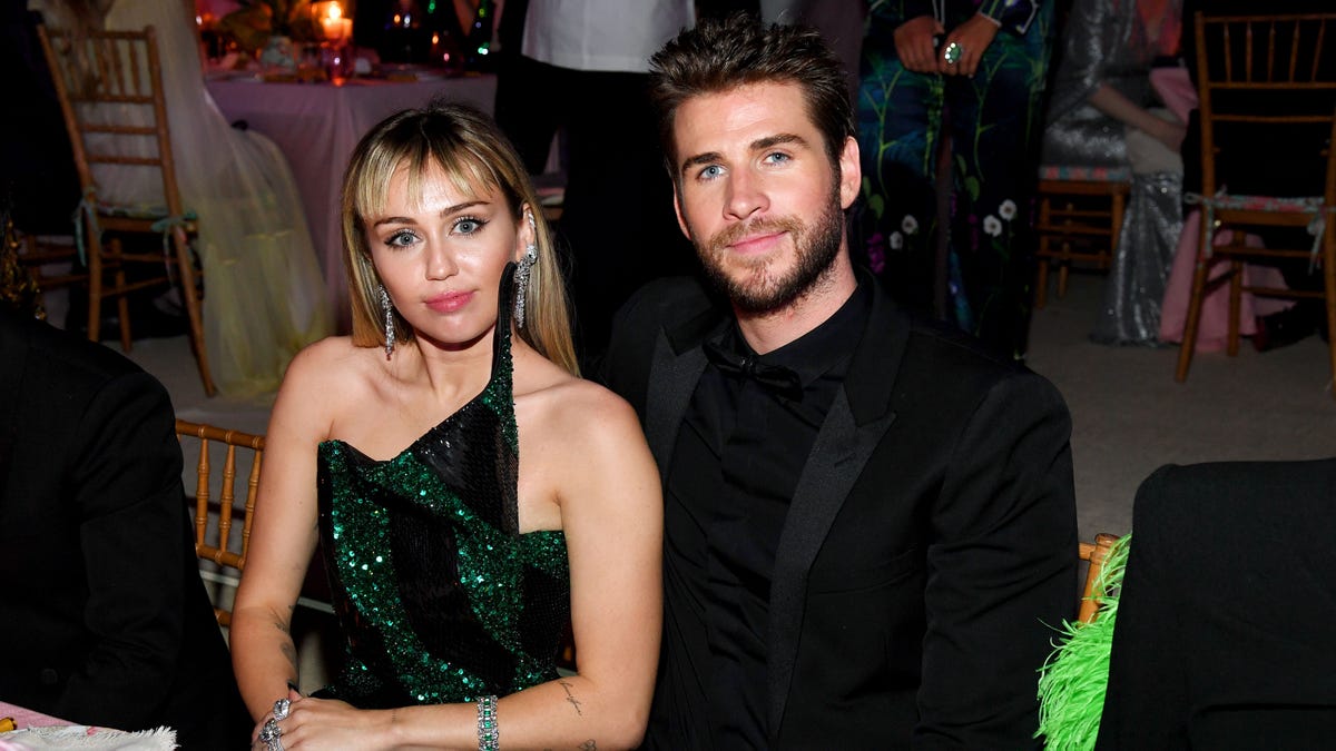 Miley Cyrus in a green and black gown sits at a table next to Liam Hemsworth in all black at The Met