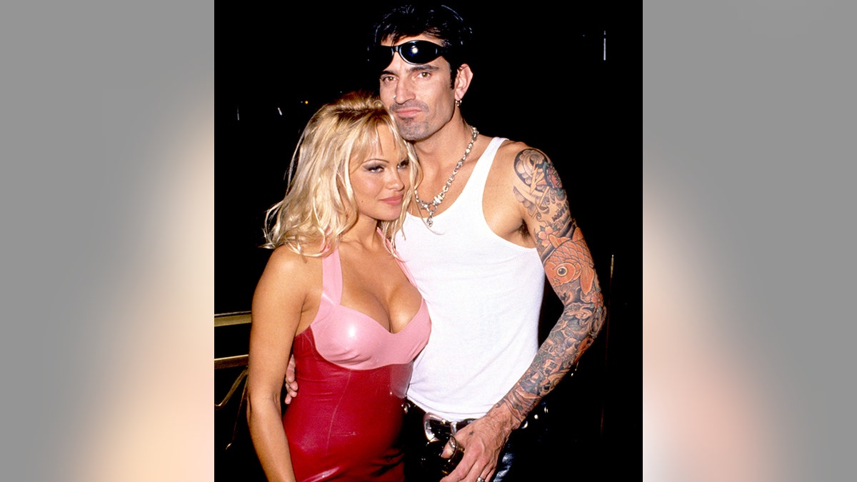 Pamela Anderson in a latex pink and red outfit smiles next to Tommy Lee in a white tank top