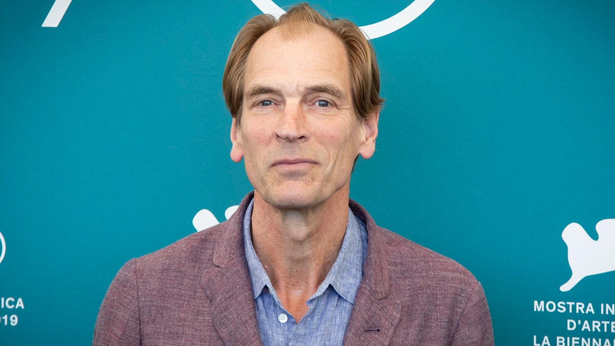 Julian Sands soft smiles on the carpet in a light burgundy jacket and blue shirt