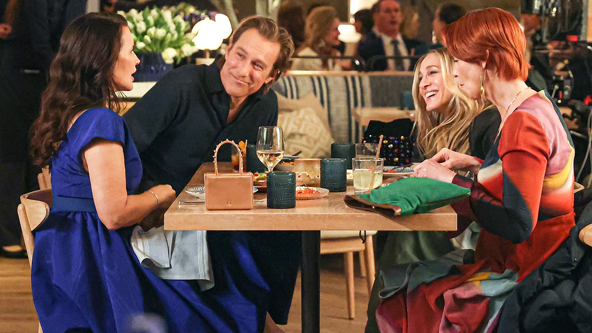 Kristin Davis (Charlotte) John Corbett (Aidan) Sarah Jessica Parker (Carrie) and Cynthia Nixon (Miranda) sit at a table and laugh while filming a scene for "And Just Like That"